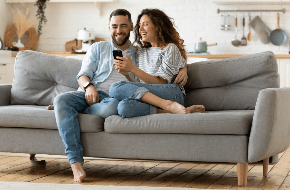 Smiling Couple sitting on a sofa, looking at a mobile screen and smiling with satisfaction
